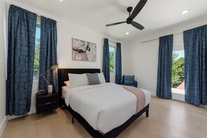 Une chambre à coucher comprenant un lit, une table de chevet, un ventilateur de plafond, une fenêtre avec des rideaux, une œuvre d’art accrochée au mur et une vue sur la verdure à l’extérieur.