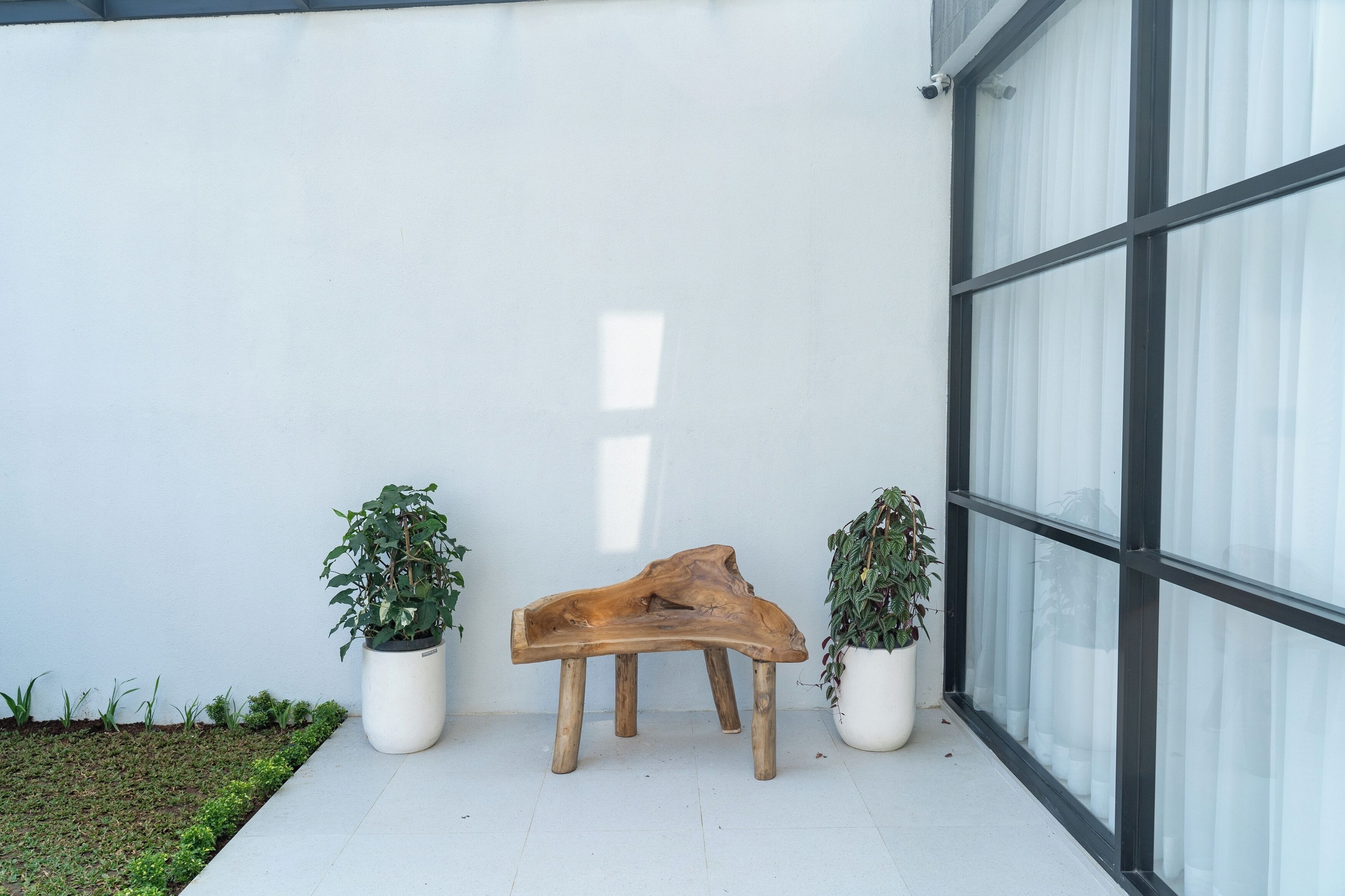 A wooden bench with two potted plants on either side, positioned against a white wall with a large window.