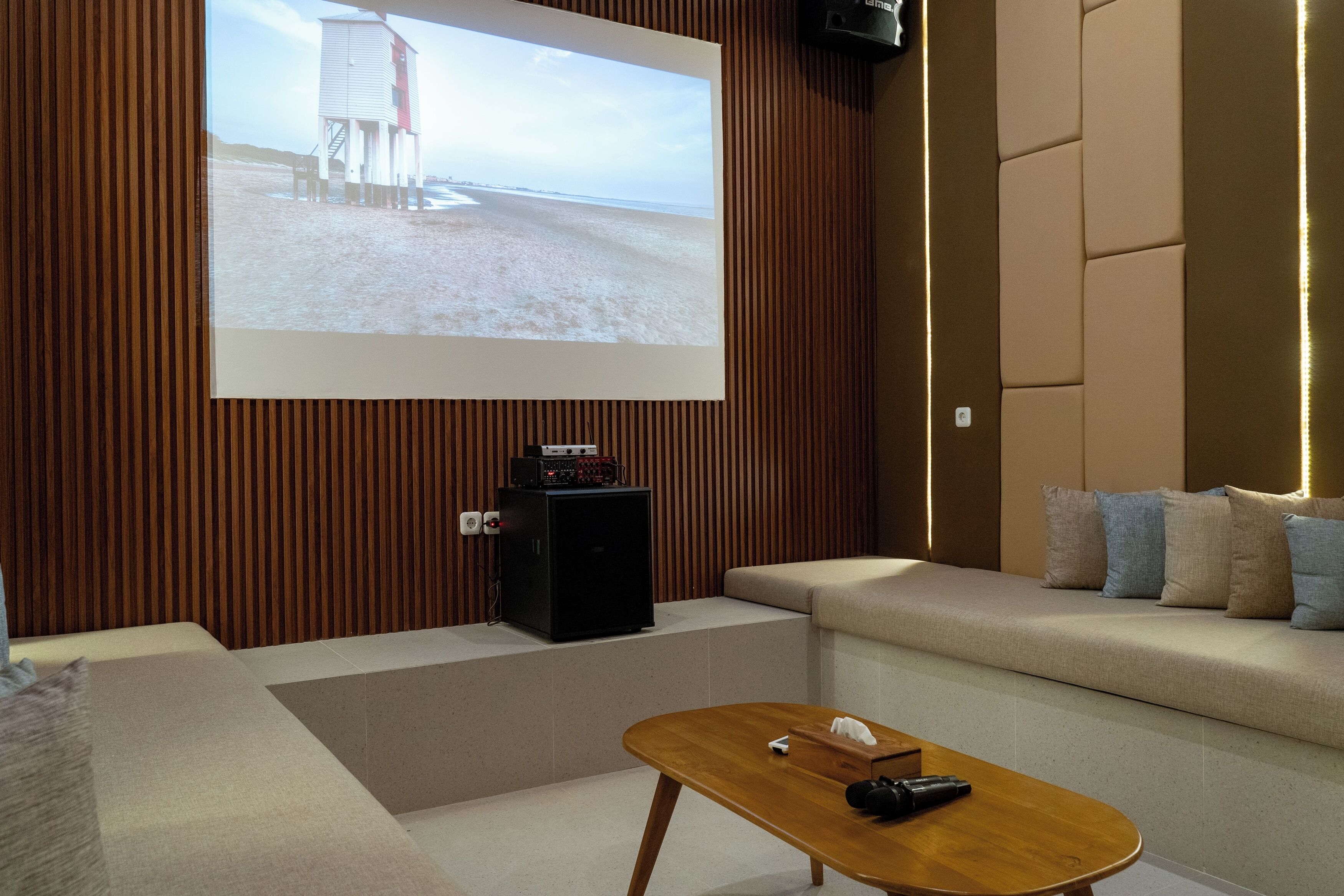 Una sala de estar moderna con un televisor de pantalla plana grande montado en una pared de madera, un altavoz negro pequeño, una mesa de centro de madera y un sofá beige con cojines.