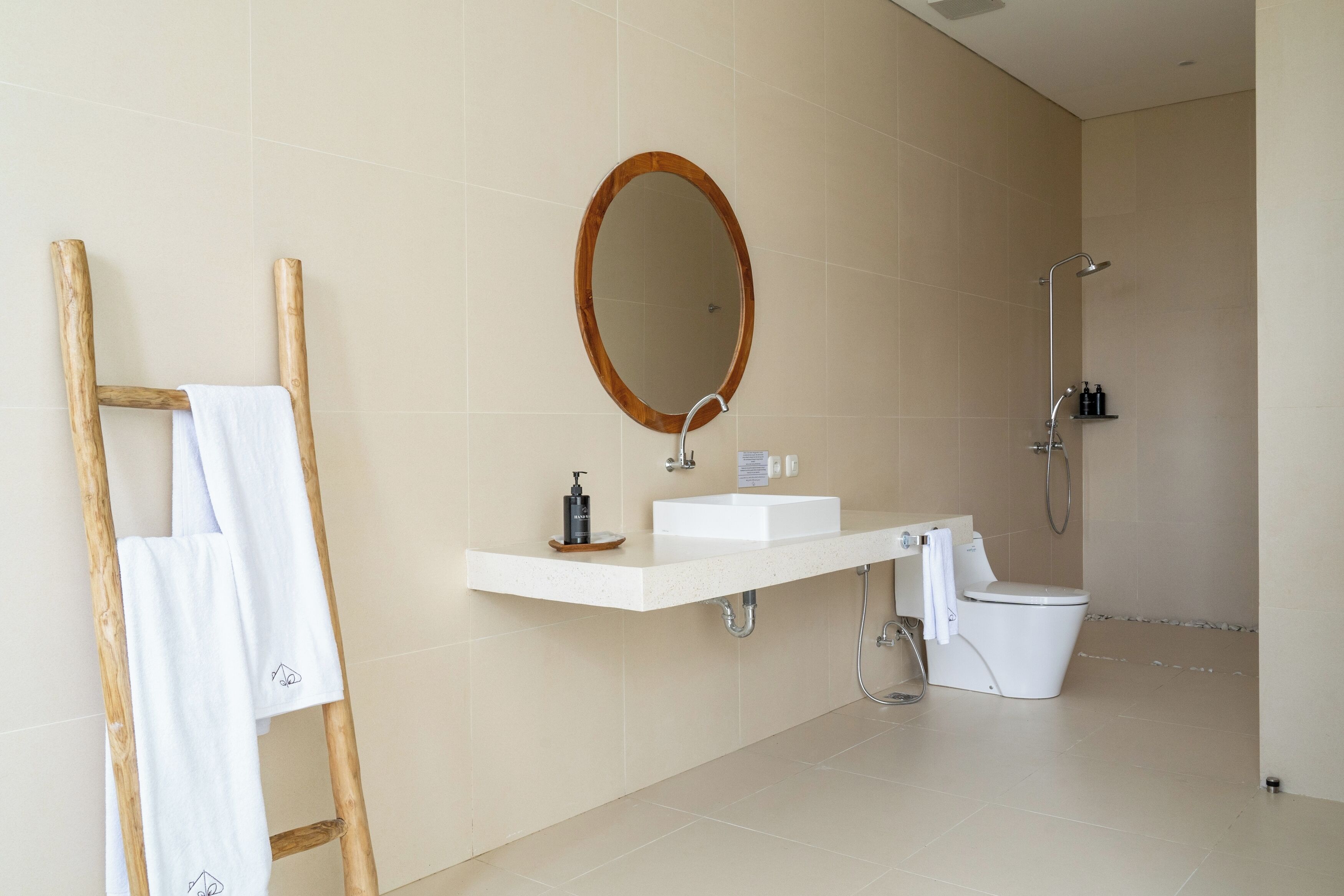 Un bagno moderno con un portasciugamani in legno, uno specchio rotondo, un lavandino, un water e una doccia.
