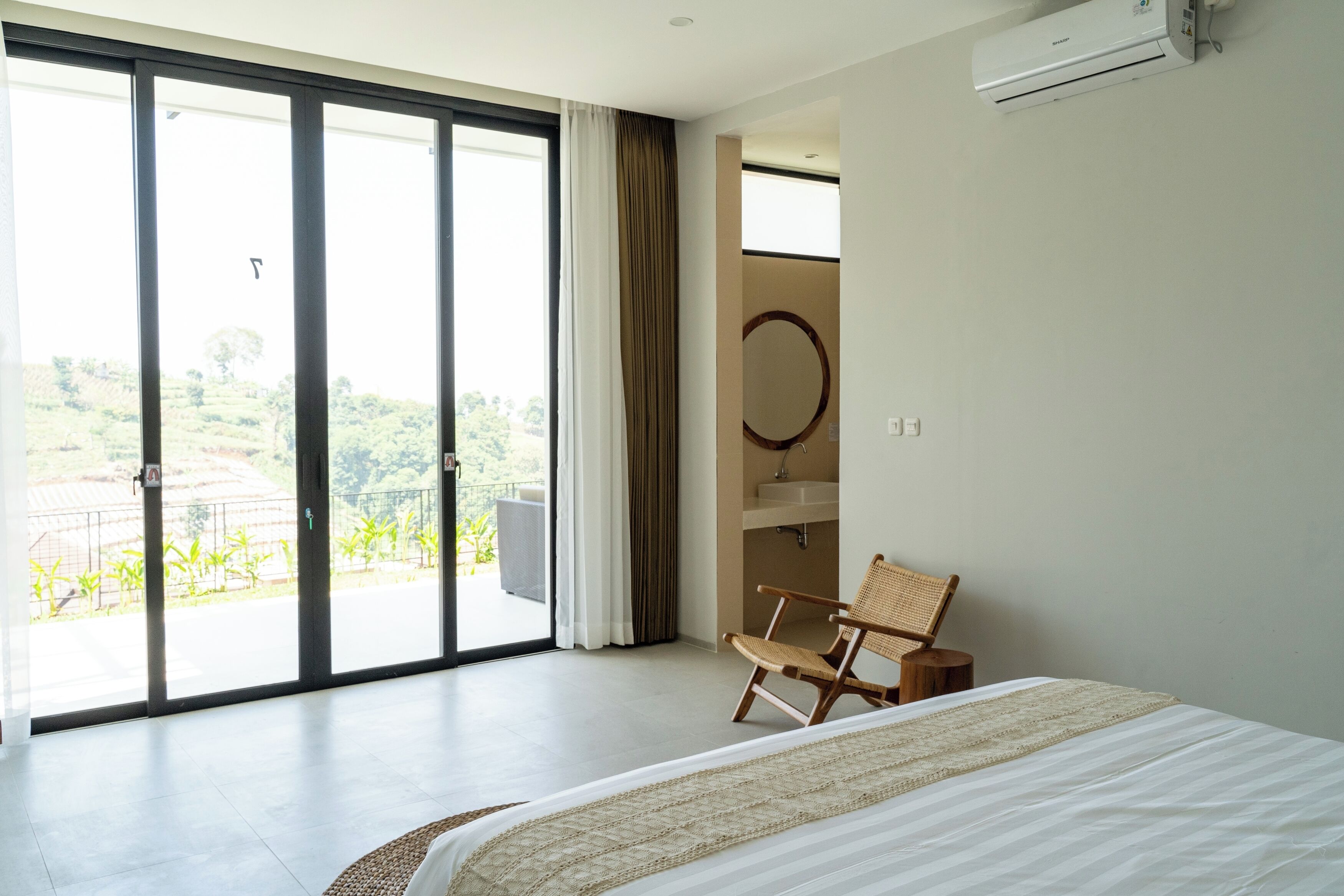 A bedroom with a large bed, a chair, a bathroom with a mirror, and a view of greenery outside.