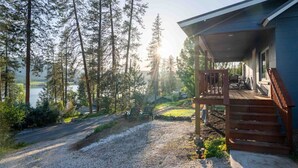 Una casa con una terrazza in legno, un vialetto sterrato e vista su un lago circondato da alberi alti.