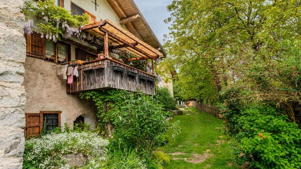 A rustic stone building with a wooden balcony, surrounded by lush greenery and a garden path.
