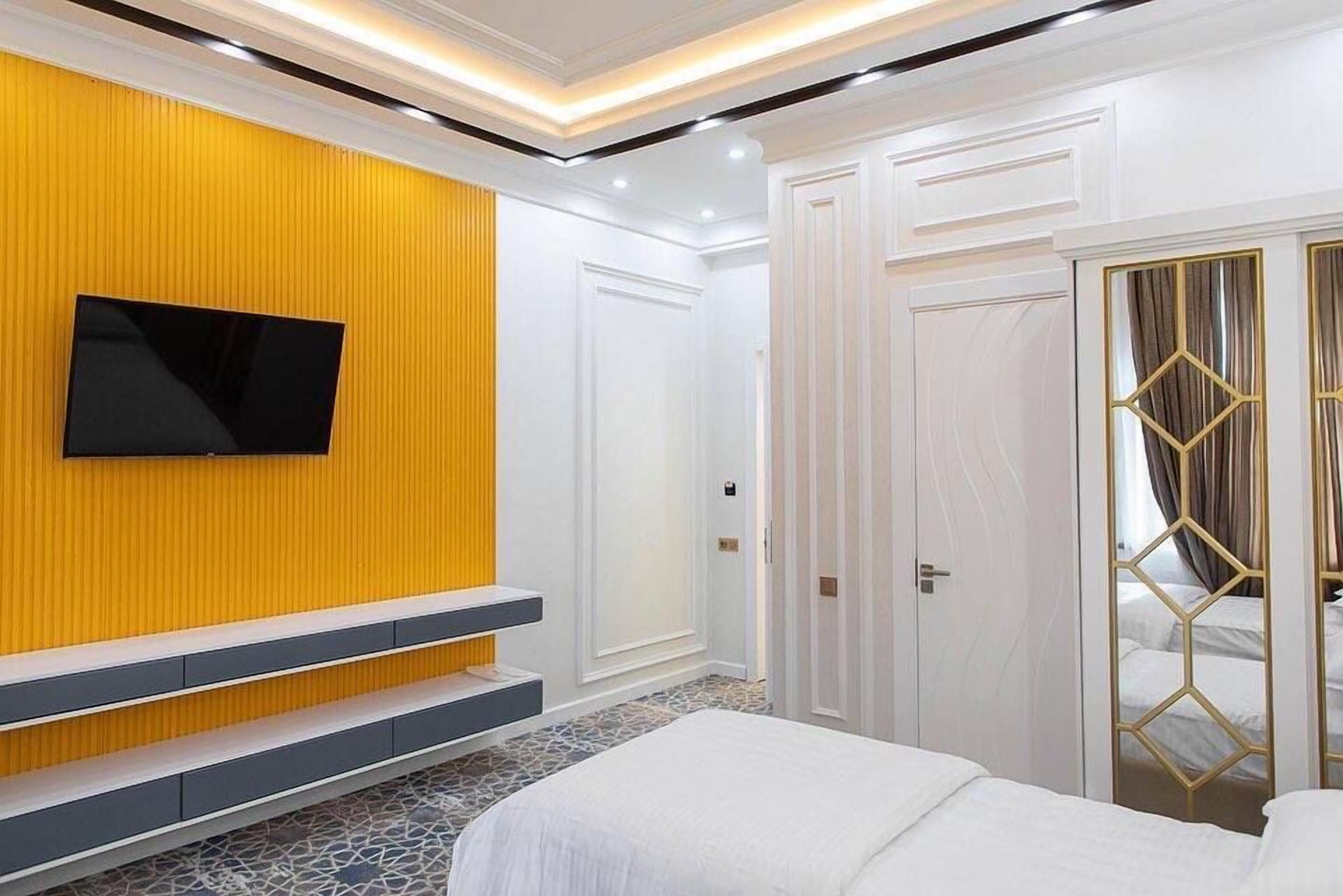 Una camera d'albergo con un televisore montato su una parete gialla, un letto con biancheria bianca e un pavimento a motivi.