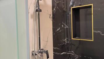 A bathroom with a glass shower enclosure, a handheld showerhead, a wall-mounted showerhead, and a marble-tiled wall.