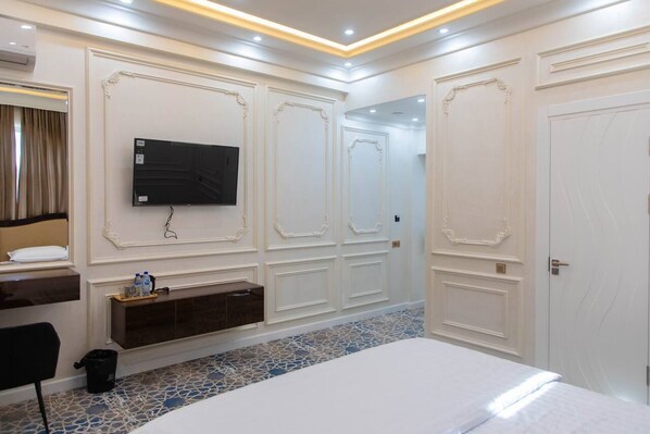 A hotel room with a large bed, a flat-screen TV mounted on the wall, a wooden bench, and a tiled floor. - Ardani HOTEL (Samarkand)
