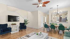 Una sala moderna con televisor de pantalla plana, sillones azul oscuro, una zona de comedor con sillas verdes, piso de madera y ventilador de techo.