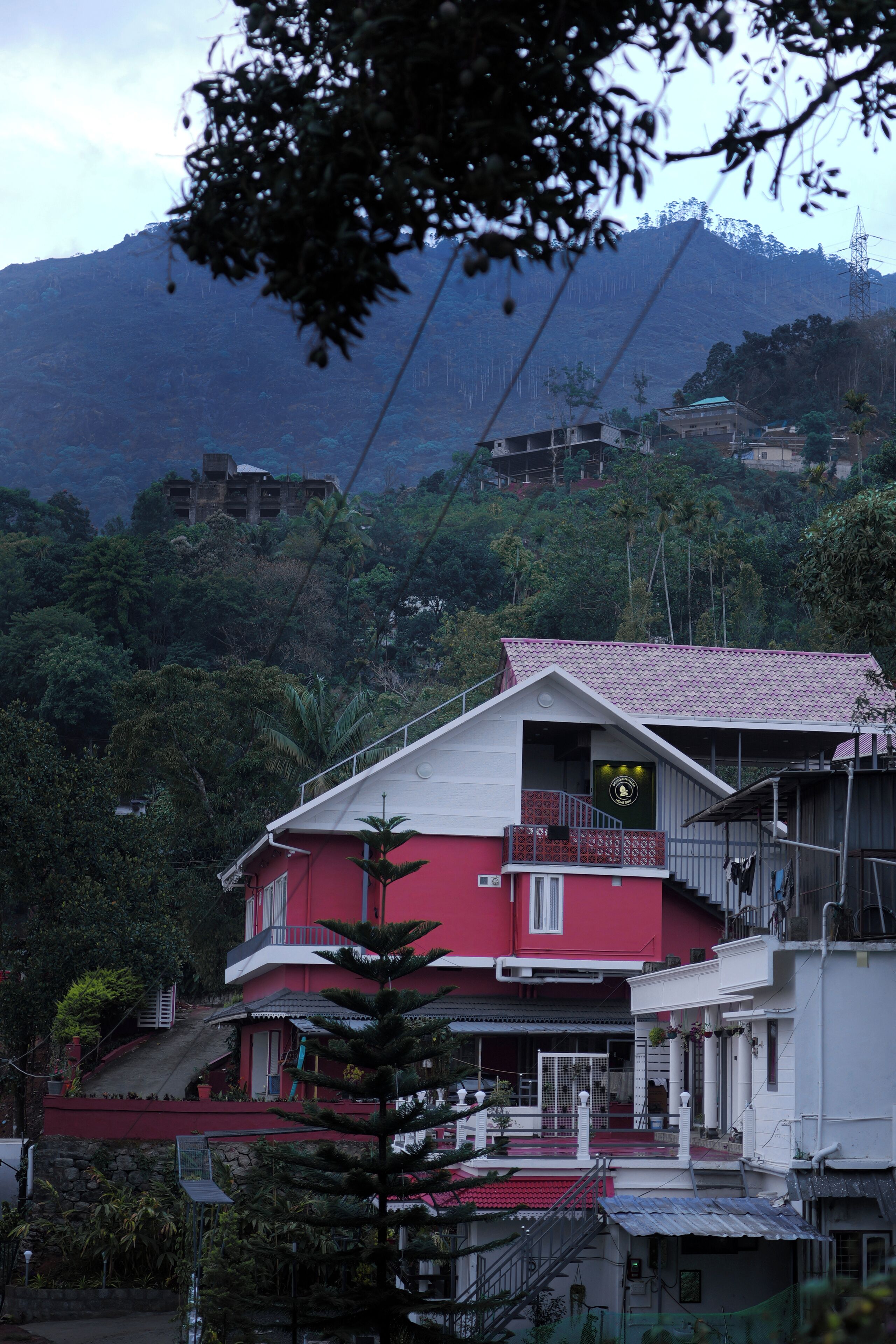 A red and white building with a balcony, surrounded by lush greenery and a mountain in the background.