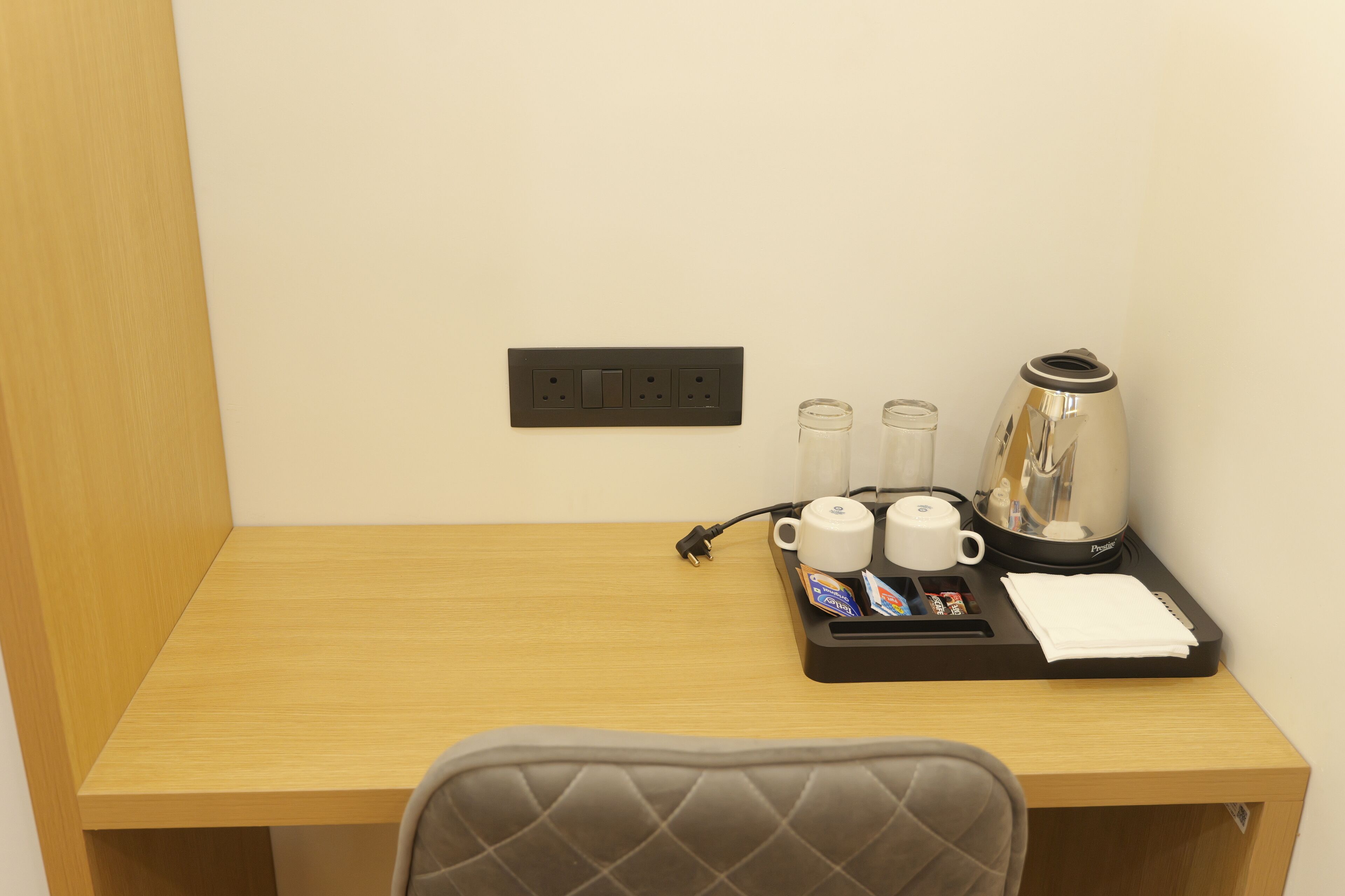 A hotel room with a wooden desk, a black tray with a coffee maker, two cups, and a power outlet.