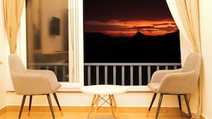 Ein Zimmer mit zwei Stühlen und einem kleinen Tisch am Fenster mit Blick auf einen Sonnenuntergang über den Bergen.