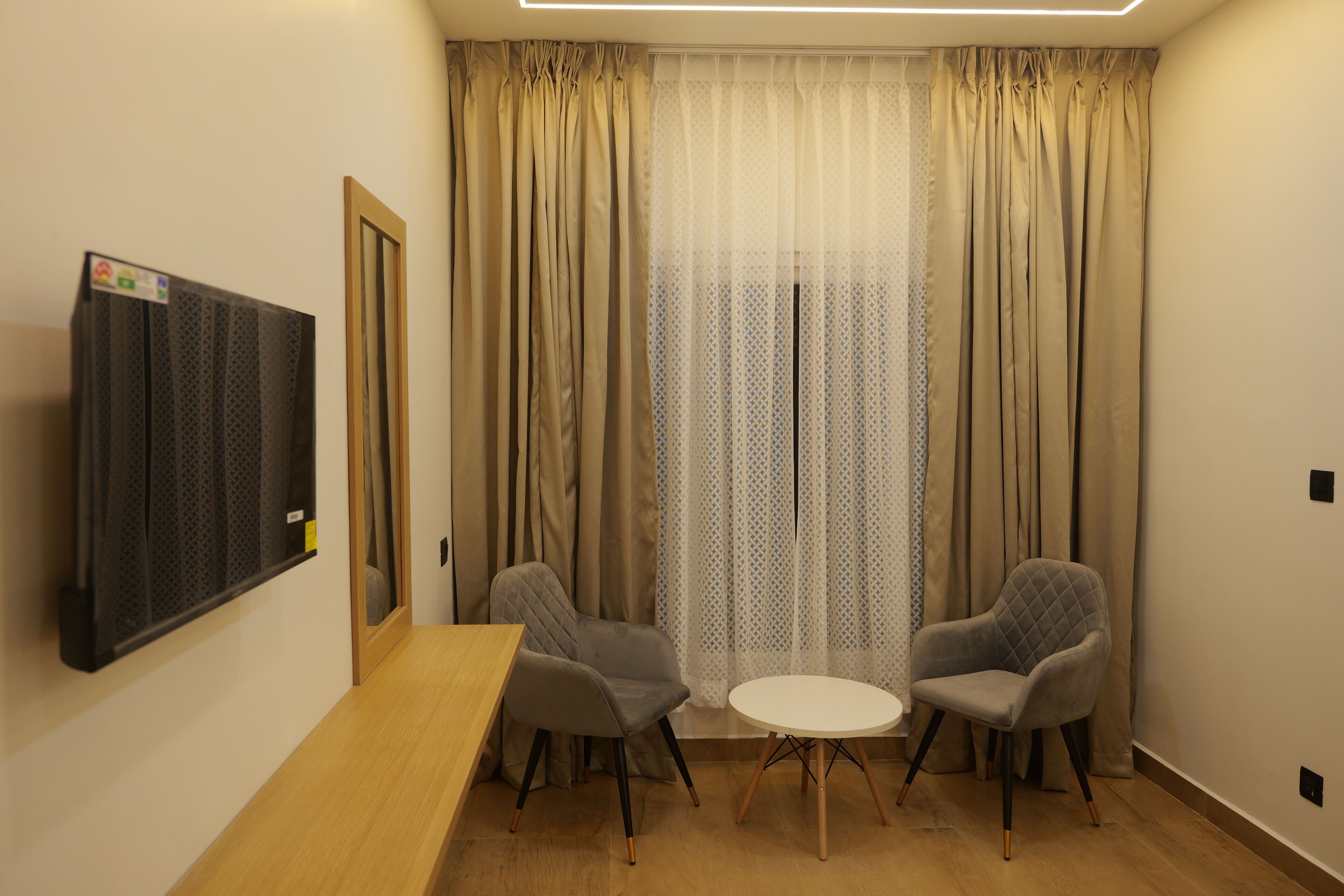 Una habitación con una mesa de madera, dos sillones grises, una mesita blanca, un televisor montado en la pared y una ventana con cortinas color beige.