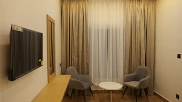 Una habitación con una mesa de madera, dos sillones grises, una mesita blanca, un televisor montado en la pared y una ventana con cortinas color beige.