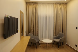 A room with a wooden table, two grey armchairs, a small white table, a TV mounted on the wall, and a window with beige curtains. - Bougainvillea Homestay Munnar (Devikolam)