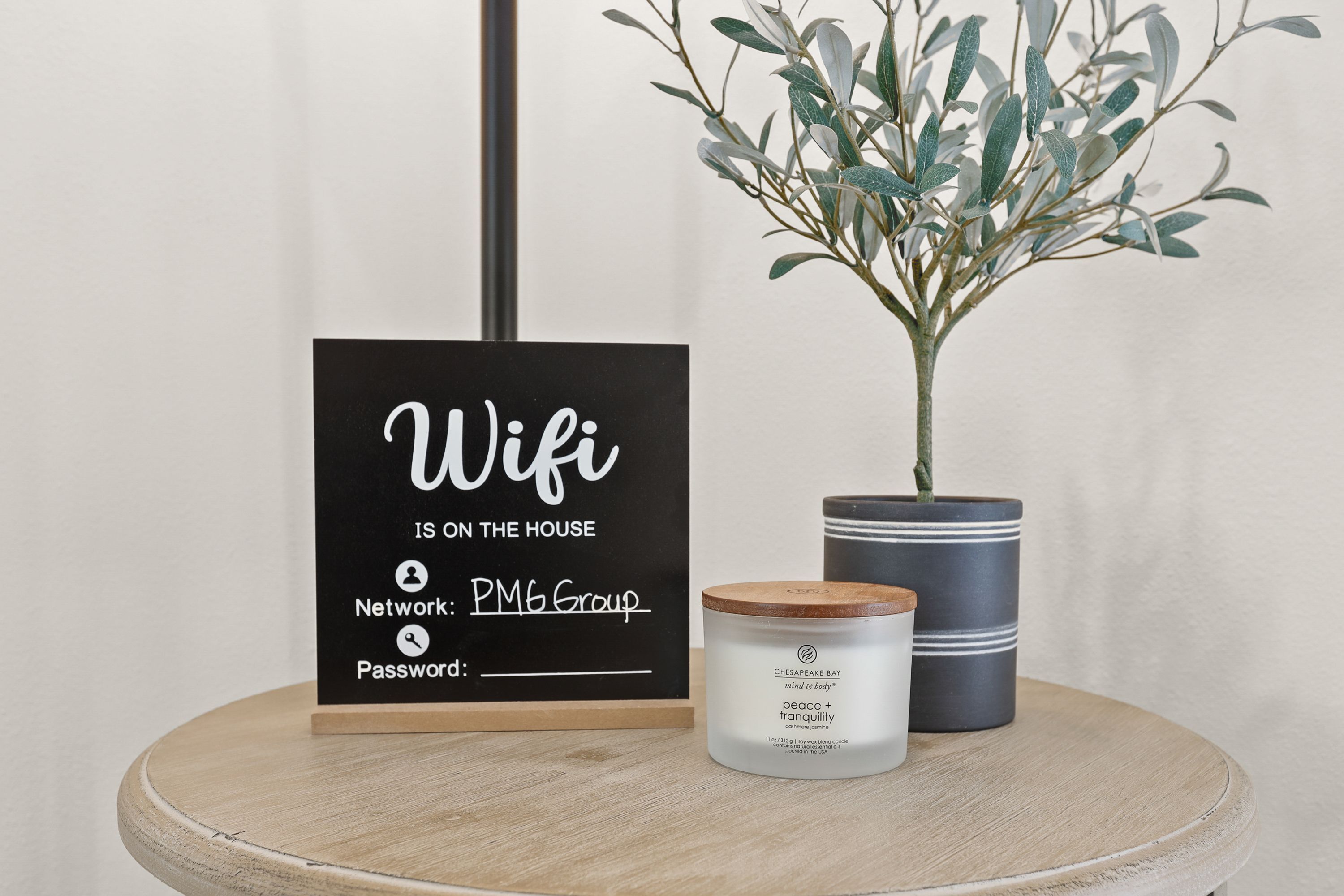 Sinal indicando 'Wifi é cortesia', 'Rede: PM6 Group' e um espaço reservado para a senha, uma vela e um vaso de planta sobre uma mesa redonda.