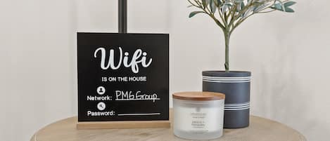 A sign indicating 'Wifi is on the house' with 'Network: PM6 Group' and a placeholder for a password, a candle, and a potted plant on a round table.