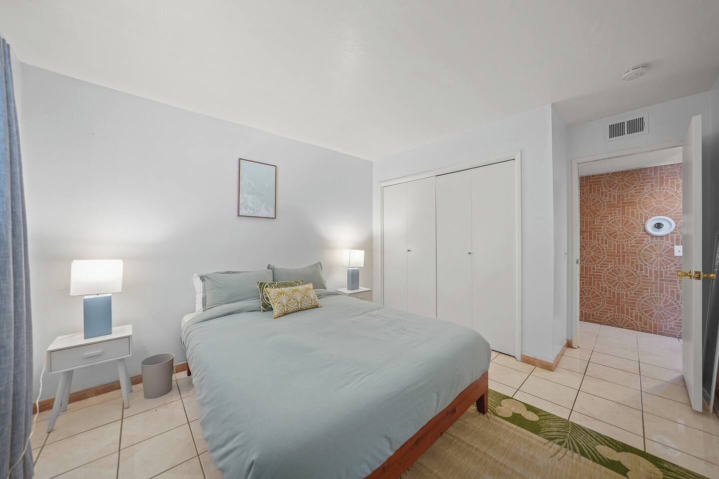 Een slaapkamer met een bed, twee nachtkastjes met lampen, een prullenbak, een deur met een patroon op de muur, en een plafondlamp.