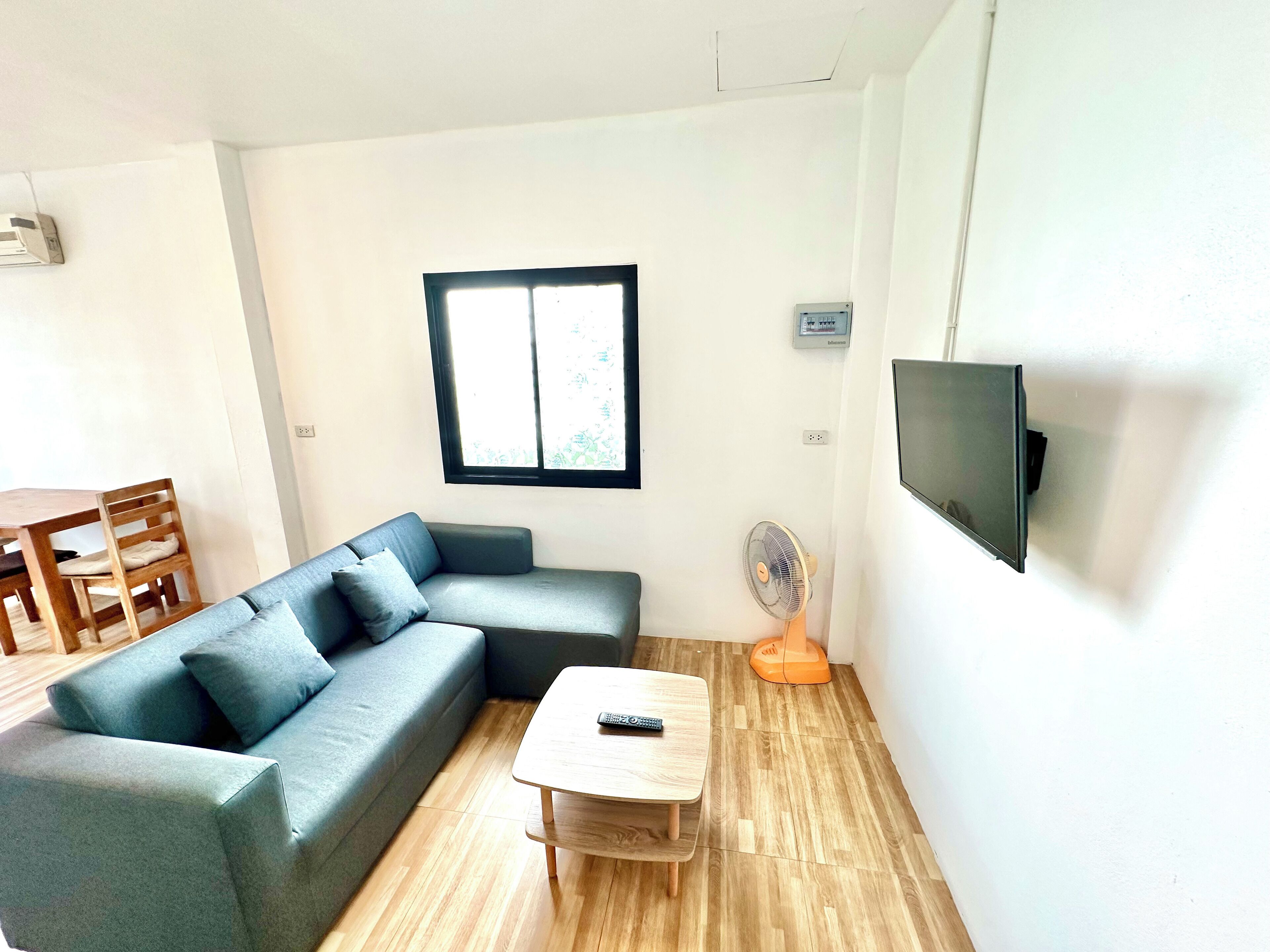 A living room with a grey sofa, a wooden coffee table, a TV mounted on the wall, and a window with blinds.