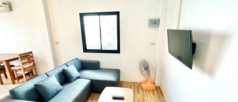 A living room with a grey sofa, a wooden coffee table, a TV mounted on the wall, and a window with blinds.