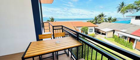 A balcony with wooden furniture, a clear view of a beach, and a red-roofed building in the background.