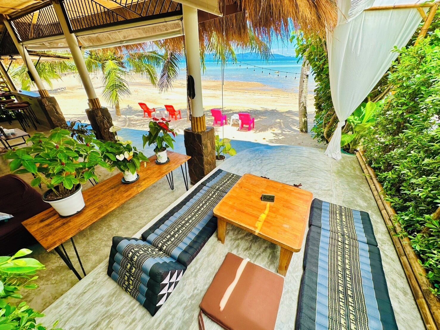 Ein Sitzbereich am Strand mit einem Holztisch, einer Couch und einer Bank. Auf dem Tisch stehen Blumentöpfe und es bietet sich ein Blick auf den Strand mit Palmen und Stühlen.