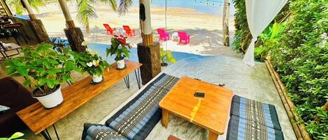Ein Sitzbereich am Strand mit einem Holztisch, einer Couch und einer Bank. Auf dem Tisch stehen Blumentöpfe und es bietet sich ein Blick auf den Strand mit Palmen und Stühlen.
