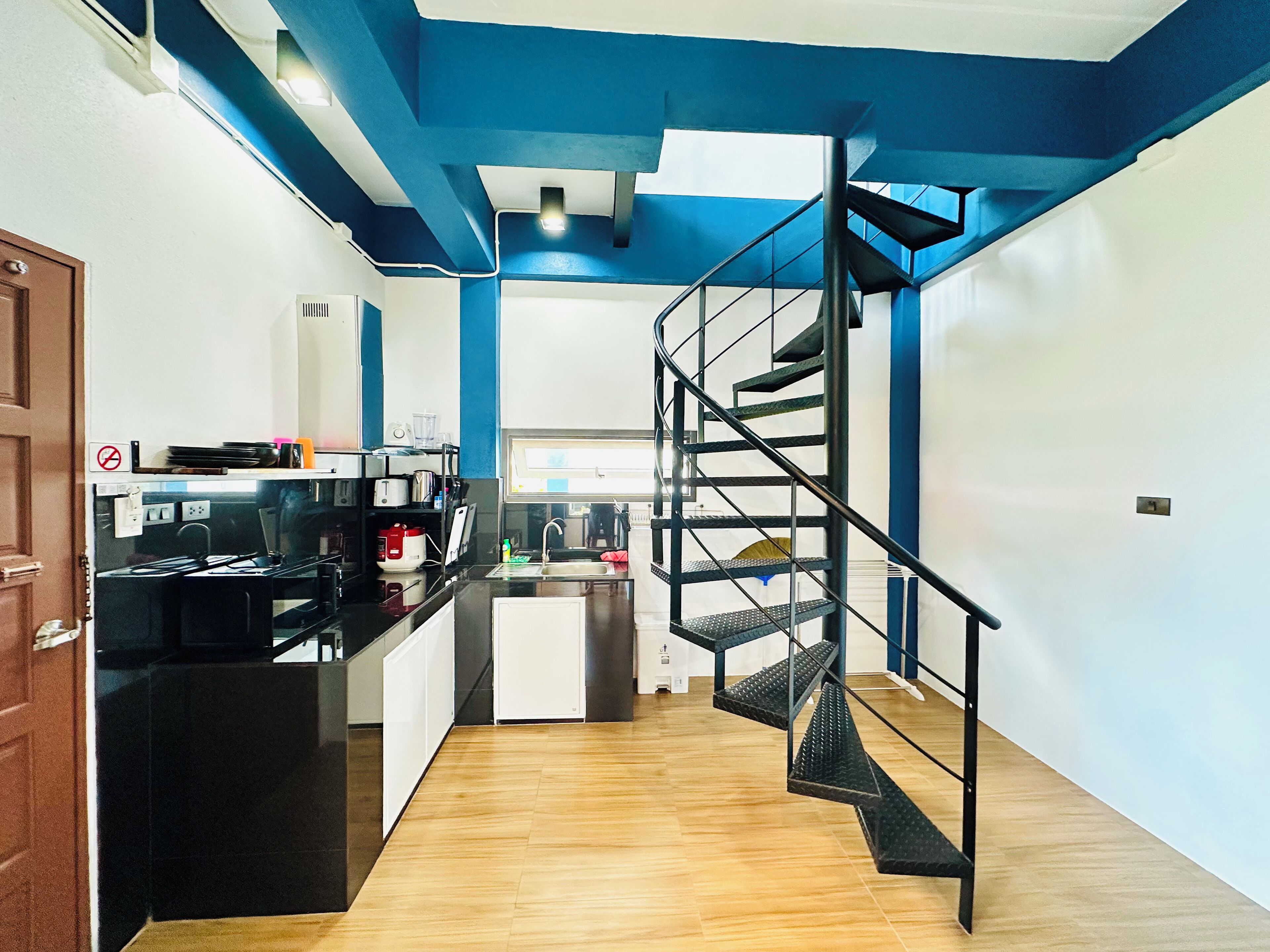 A modern kitchen with a black countertop, a built-in oven, and a microwave. There is a wooden floor and a blue accent wall. A black spiral staircase leads to the upper floor.
