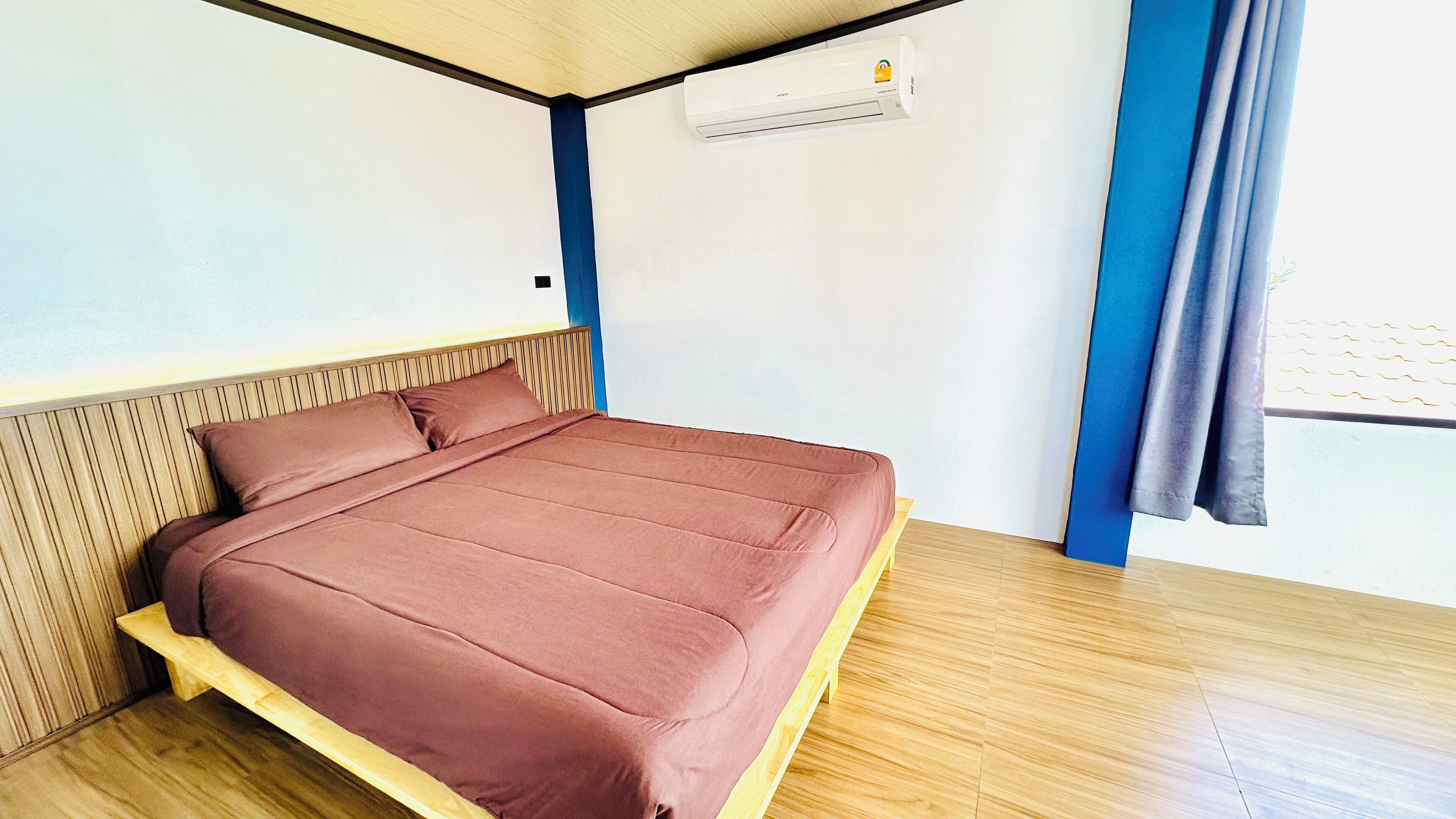 A single bed with a wooden frame and a brown mattress in a room with a sloped ceiling, a window with curtains, and an air conditioning unit on the wall.