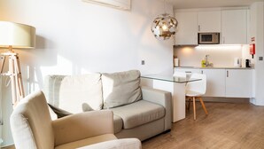A modern living room with a beige sofa, a glass dining table, and a kitchen area with white cabinets and a microwave. - Golden escape in Funchal (Funchal)