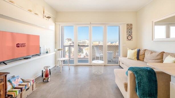 A spacious living room with a large window, a sofa, a TV, and a small table. - GuestReady - Seaview in Costa da Caparica (Almada)