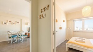 A hotel room with a bed, a window with a view, a dining area with chairs and a table, and a wall with a 'Happy Hello' sign. - GuestReady - Seaview in Costa da Caparica (Almada)