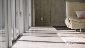 A modern interior with a glass door, a beige sofa with a pink pillow, and a striped rug on a tiled floor. - Guest House Karst (Motobu)