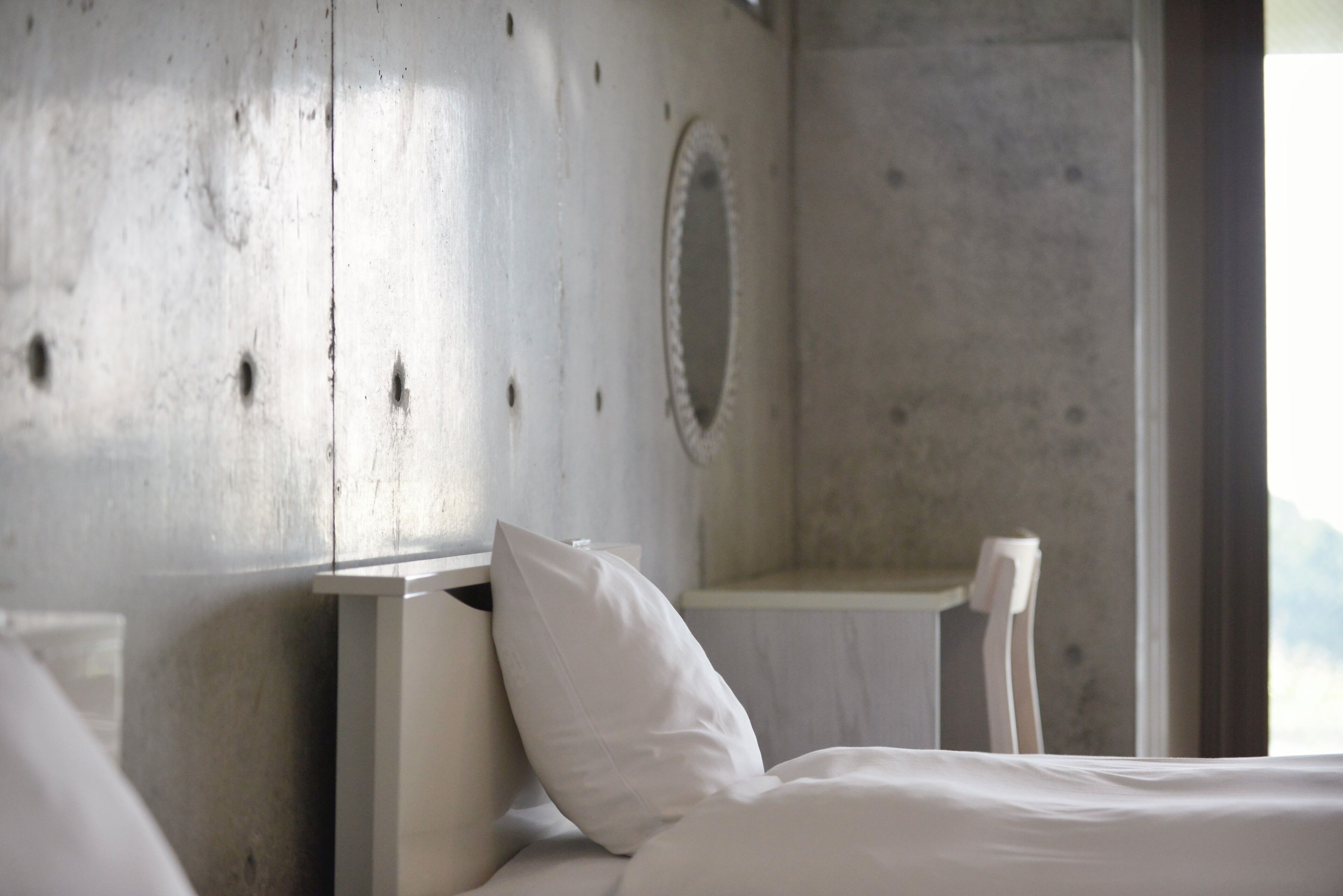 Un dormitorio con pared de concreto, una cama con ropa de cama blanca y una mesita de noche pequeña.