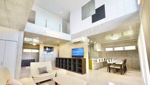 A spacious living area with a high ceiling, a flat-screen TV mounted on a stone wall, a dining area with a long table and chairs, and a kitchen in the background. - Guest House Karst (Motobu)