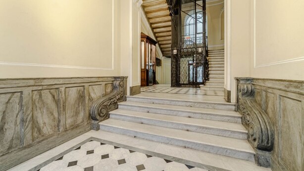 Un magnifique escalier en marbre, avec des marches et des rampes richement décorées, menant à une pièce dotée d’une grande fenêtre et d’une grille en fer forgé ornementale.