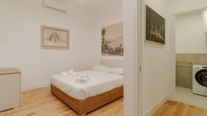 Une chambre avec un lit en bois, du linge de lit blanc, une petite table de chevet, un climatiseur fixé au mur et un lave-linge dans la pièce adjacente.