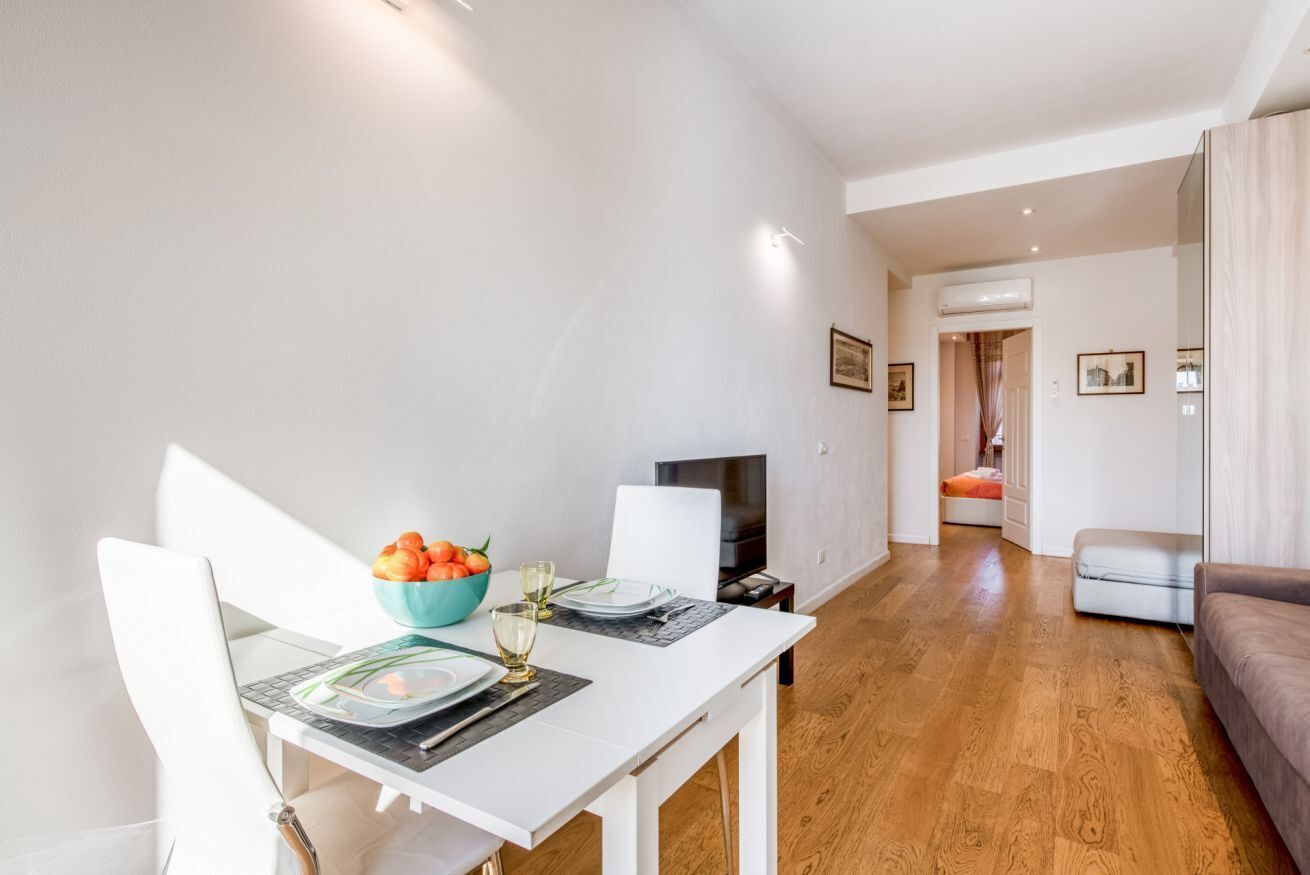 A dining area with a white table set for two, a bowl of fruit, and a television in the background.