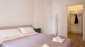 A neatly made bed with a pink bedspread and white pillows, a standing lamp, a white towel rack with clothes, and a wooden floor. - GuestHost - Cozy 55 sqm apartment, for 4 people, located on the ground floor of a modern building. The property is located in a strategic area of ​​Piombino, an ideal starting point for exploring the city and the Tuscan coast. A few minutes from the  (Piombino)
