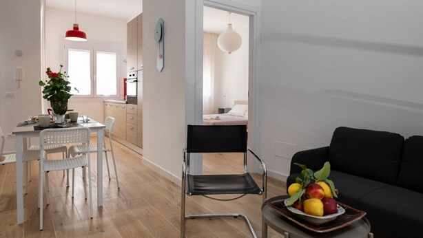 A modern living room with a black sofa, a dining area with a white table and chairs, and a kitchen visible in the background. - GuestHost - Cozy 55 sqm apartment, for 4 people, located on the ground floor of a modern building. The property is located in a strategic area of ​​Piombino, an ideal starting point for exploring the city and the Tuscan coast. A few minutes from the  (Piombino)