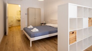 A bedroom with a bed, a wardrobe, a hanging light fixture, and a white shelving unit. - GuestHost - Cozy 40 sqm studio, for 4 people, located on the ground floor of a modern building. The property is located in a strategic area of ​​Piombino, an ideal starting point for exploring the city and the Tuscan coast. A few minutes from the his (Piombino)