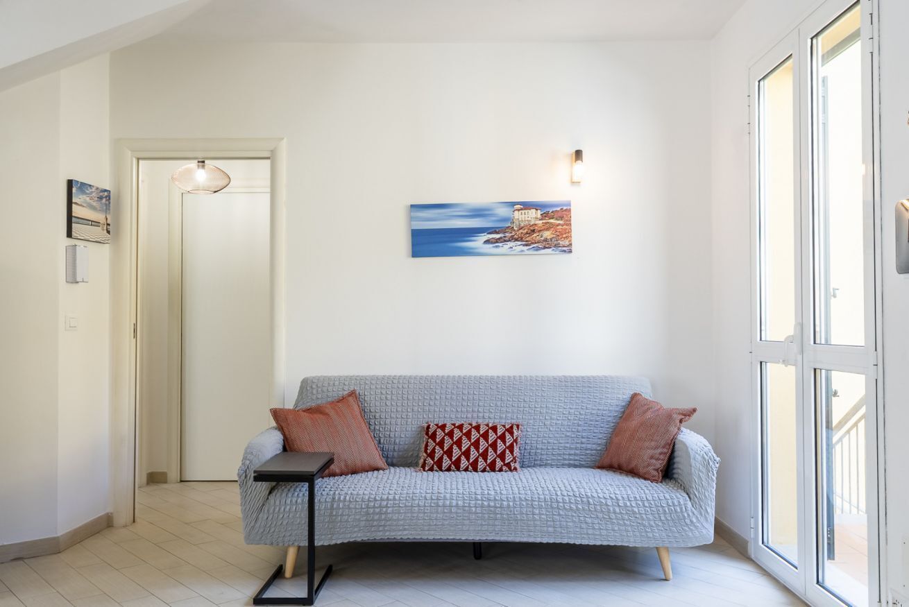 Una sala moderna con un sofá gris con textura, una mesita auxiliar, un cuadro de una escena costera y una puerta que da a otra habitación.