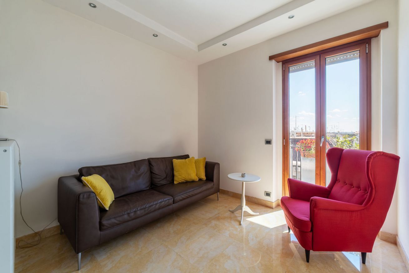 Ein modernes Wohnzimmer mit einem braunen Ledersofa, einem roten Sessel, einem kleinen weißen Tisch und einer Glastür zum Balkon.
