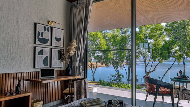 Un intérieur moderne avec une grande fenêtre donnant sur un lac, des meubles en bois et des œuvres d’art encadrées accrochées au mur.