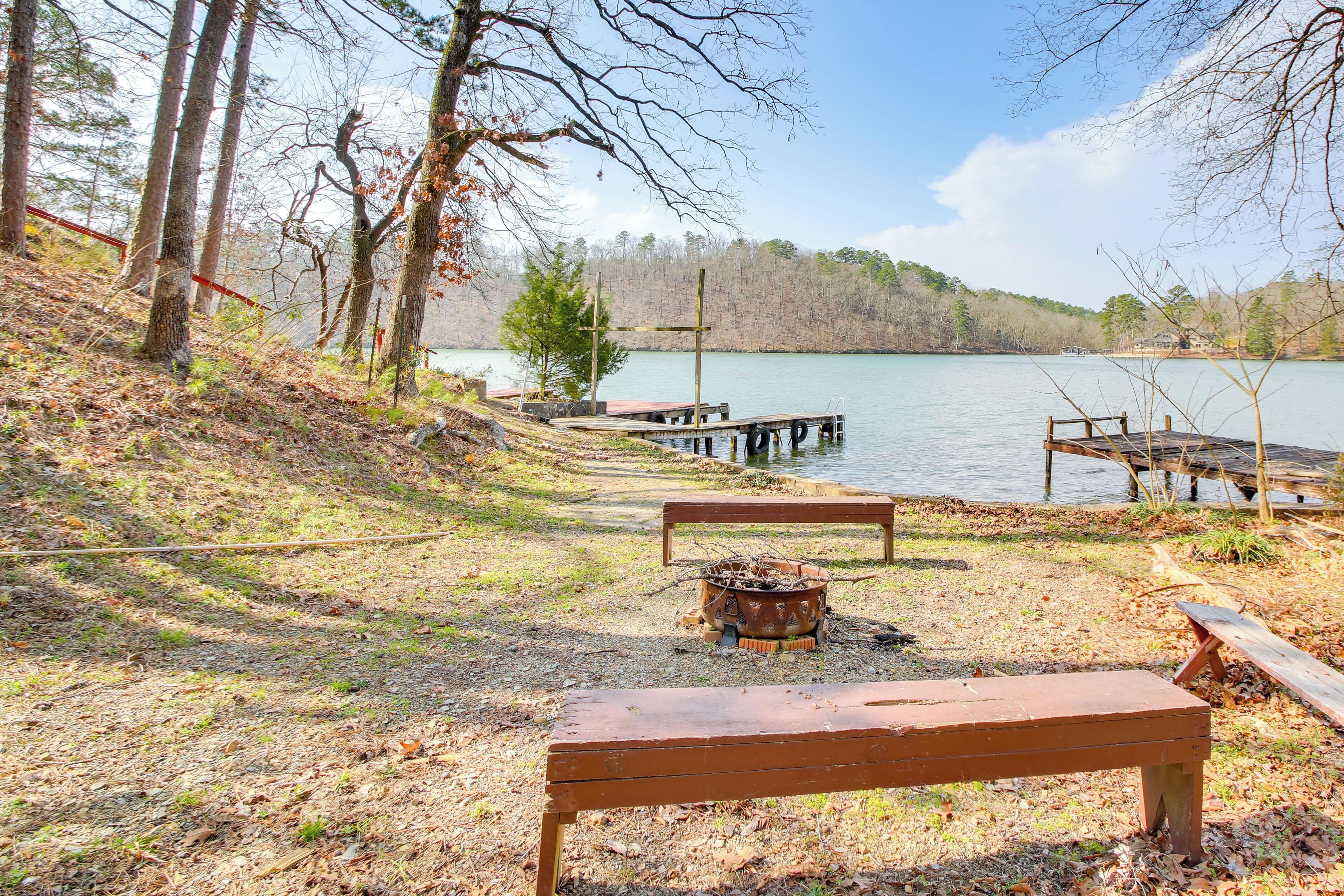 A lakeside park with wooden benches, a fire pit, and a dock extending into the water.