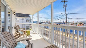 A covered porch with wooden chairs and a table, overlooking a beach area with a fence and power lines.