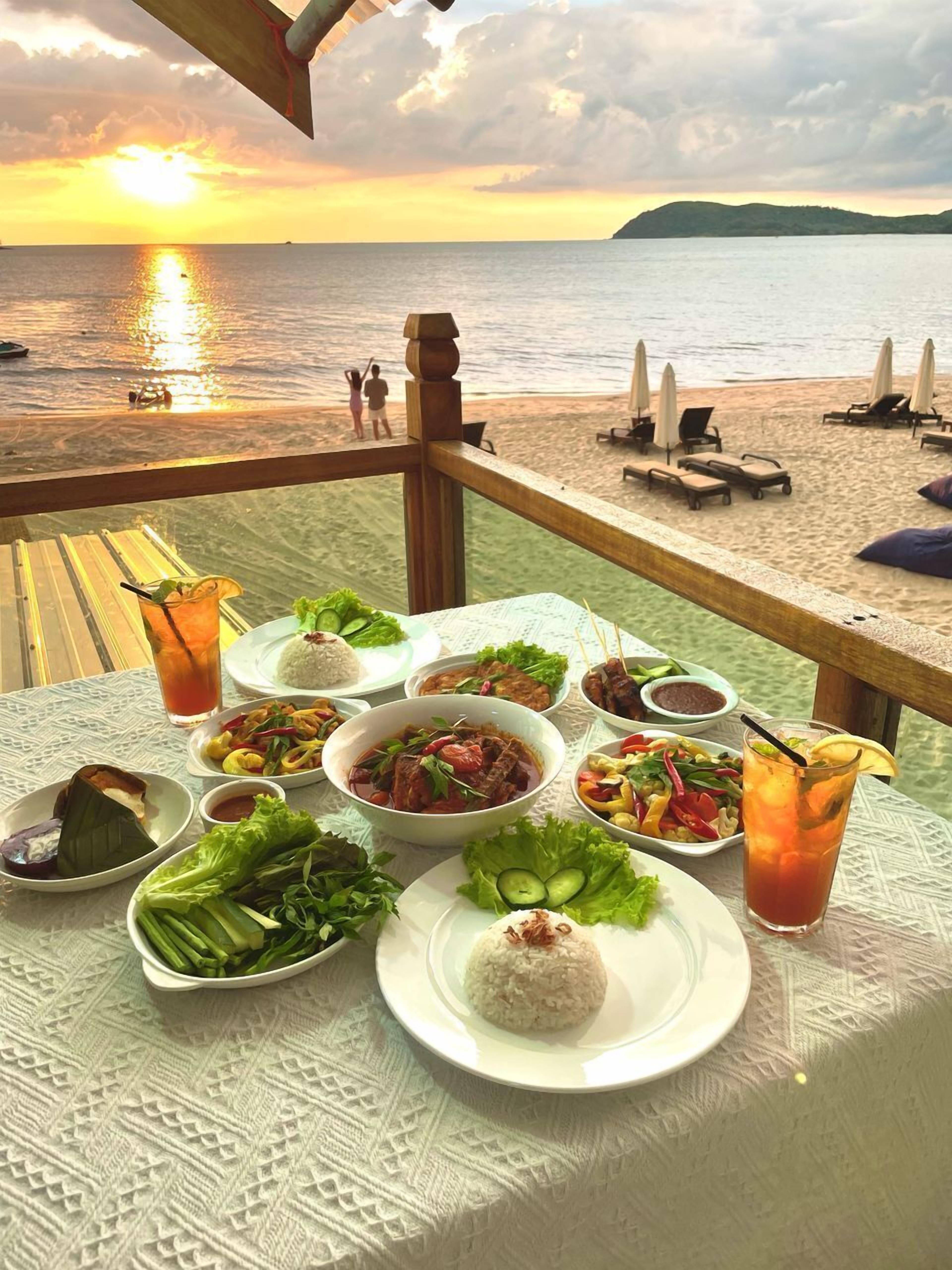 A table set with various dishes and drinks, overlooking a beach with umbrellas and lounge chairs.