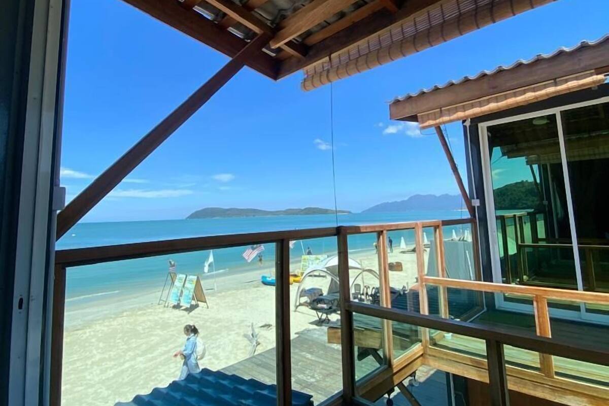 Quarto à beira-mar com amplas janelas para a praia, um deck de madeira e vista para o oceano e montanhas.