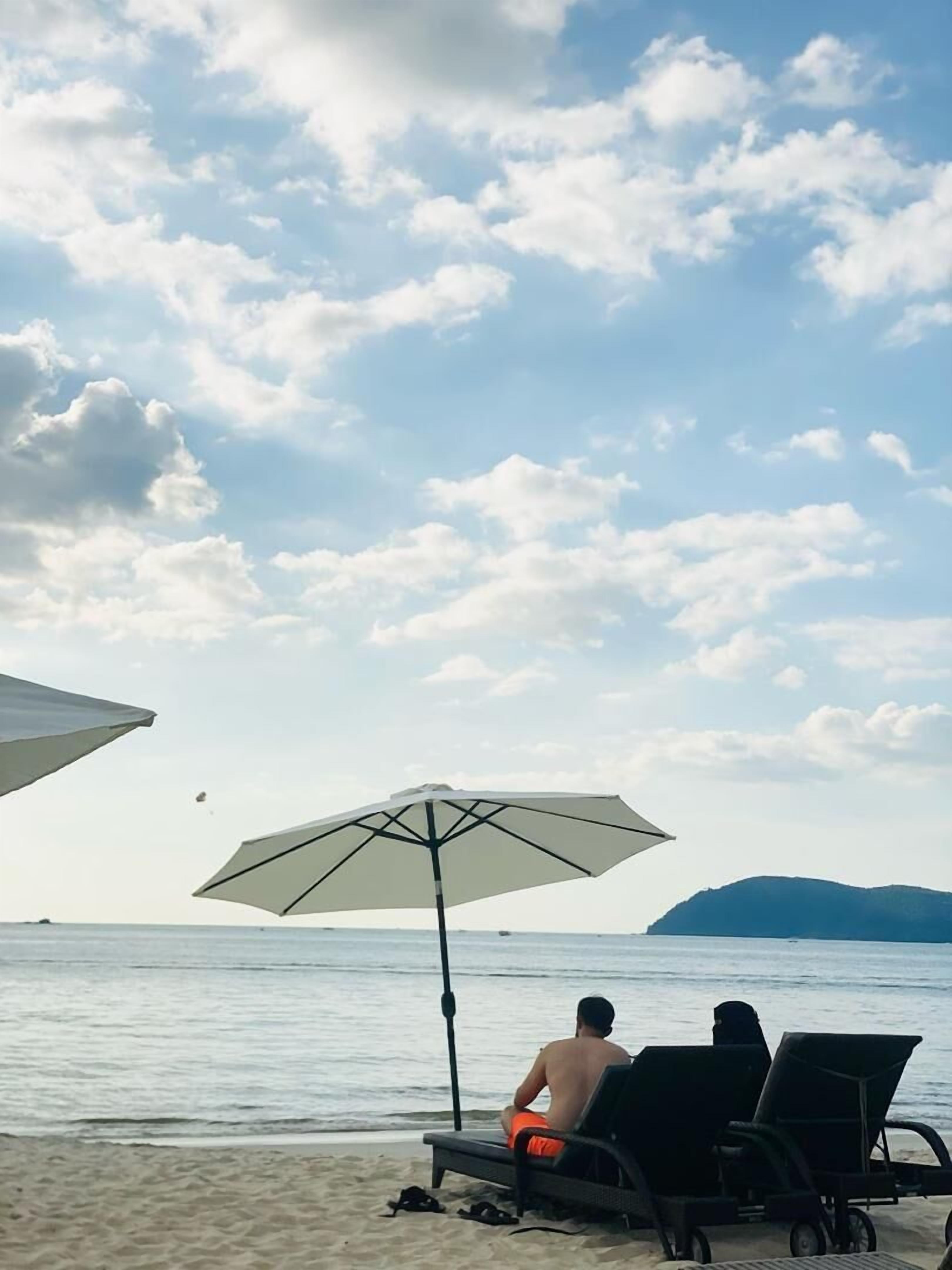 A beach scene with a person sitting on a lounge chair under an umbrella, facing the sea.