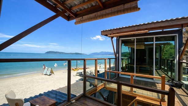 Una terraza de madera con barandilla de vidrio que da a una playa, con vista despejada al mar y montañas a lo lejos.