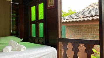 A room with a bed, a wooden floor, a balcony with a wooden railing, and a view of a brick building outside.