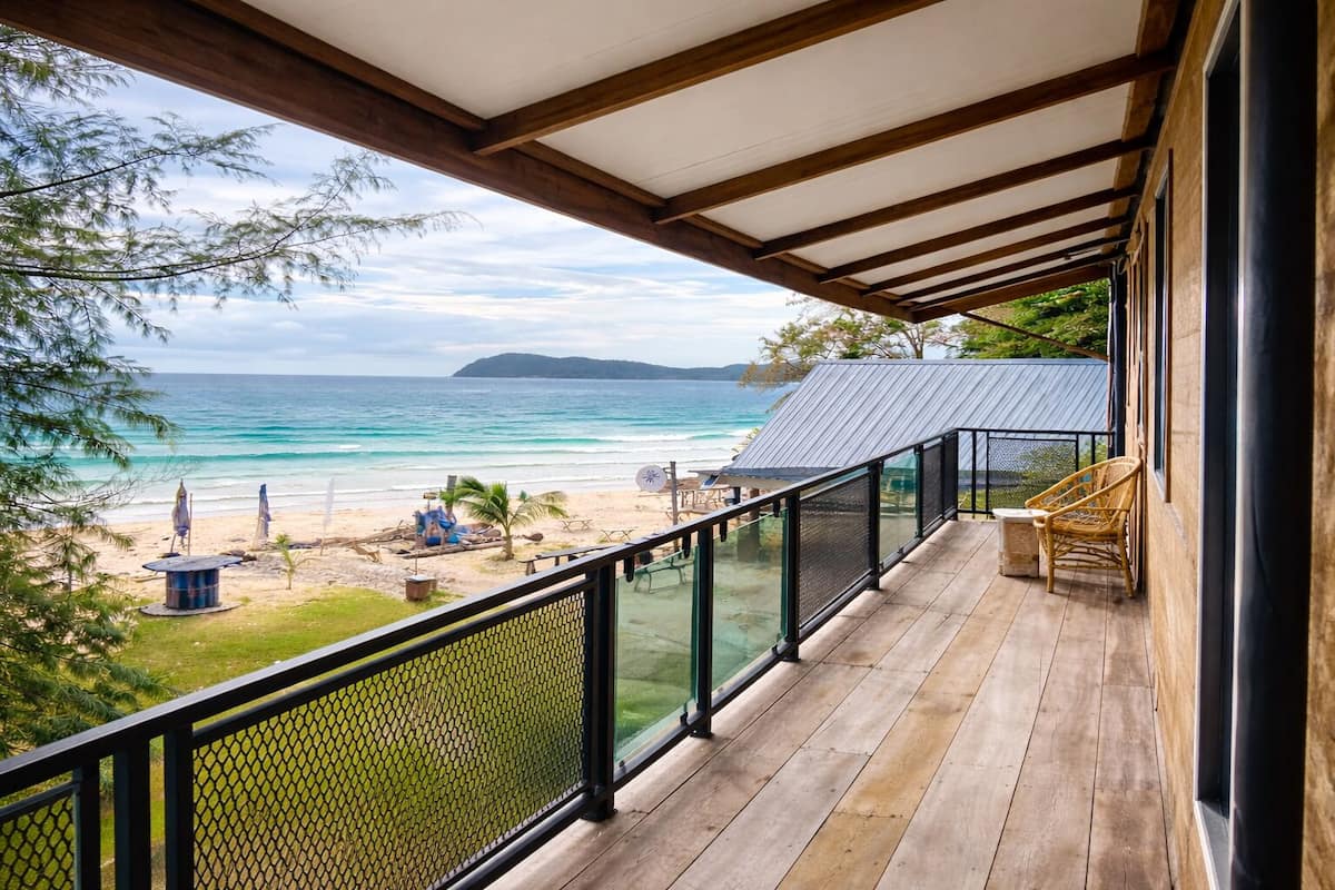 Um deck de madeira com guarda-corpo de vidro oferece vista para uma praia e para o oceano, com terra visível à distância.